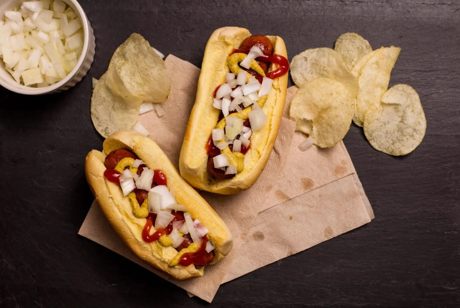 Two hot dogs topped with onions, ketchup, and mustard beside potato chips.